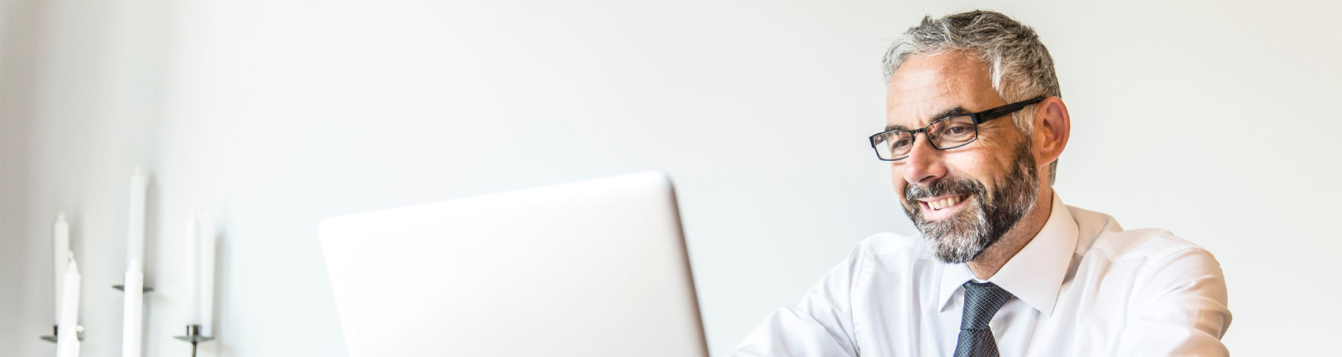 Ein lächelnder Mann mit Brille sitzt am Laptop, umgeben von Kerzen in einem modernen Büro.