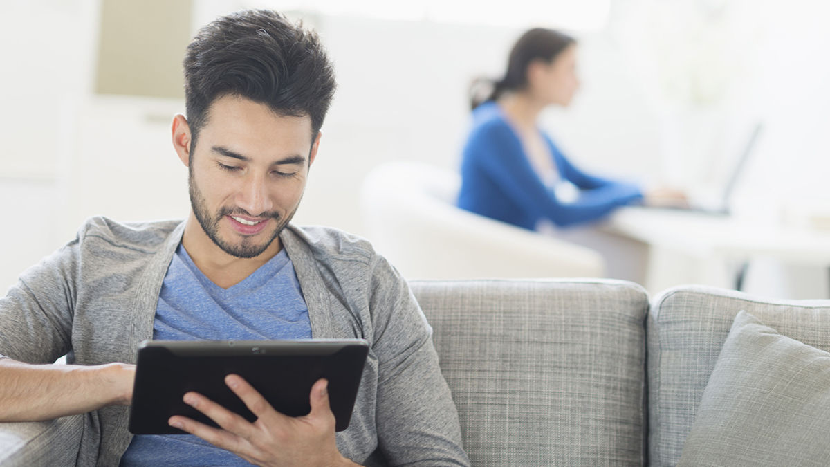 Ein Mann sitzt auf einer Couch und nutzt ein Tablet, während eine Frau im Hintergrund am Computer arbeitet.