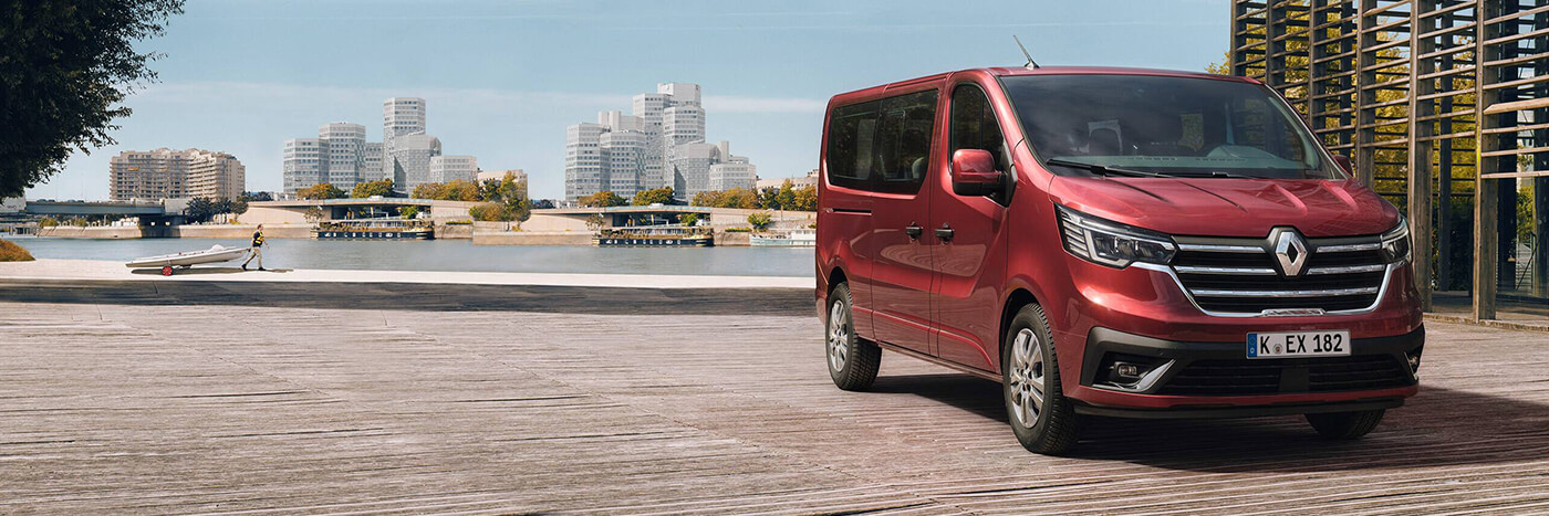 Roter Renault Van vor einer modernen Stadtlandschaft am Wasser, mit Holzsteg im Vordergrund.