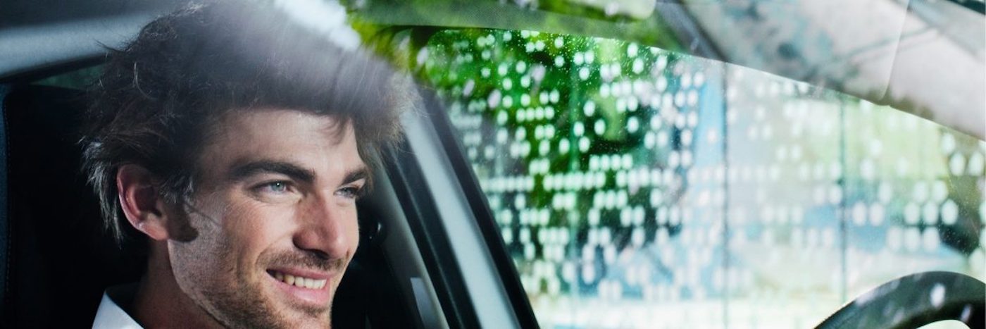 Junger Mann sitzt lächelnd im Auto, während Regen auf die Windschutzscheibe prasselt.