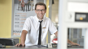 Lächelnder Mitarbeiter am Empfangstresen, trägt Hemd und Krawatte, im Hintergrund Werbung für Renault Dienstleistungen.