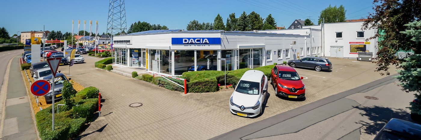 Dacia Autohaus mit mehreren Fahrzeugen auf dem Parkplatz und starker Sonneneinstrahlung, umgeben von Bäumen.