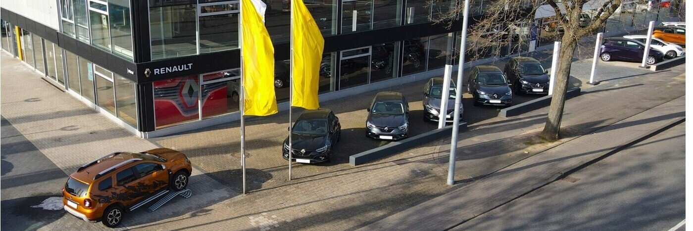 Renault Autohaus mit mehreren Autos in verschiedenen Farben und großen gelben Fahnen im Vordergrund.