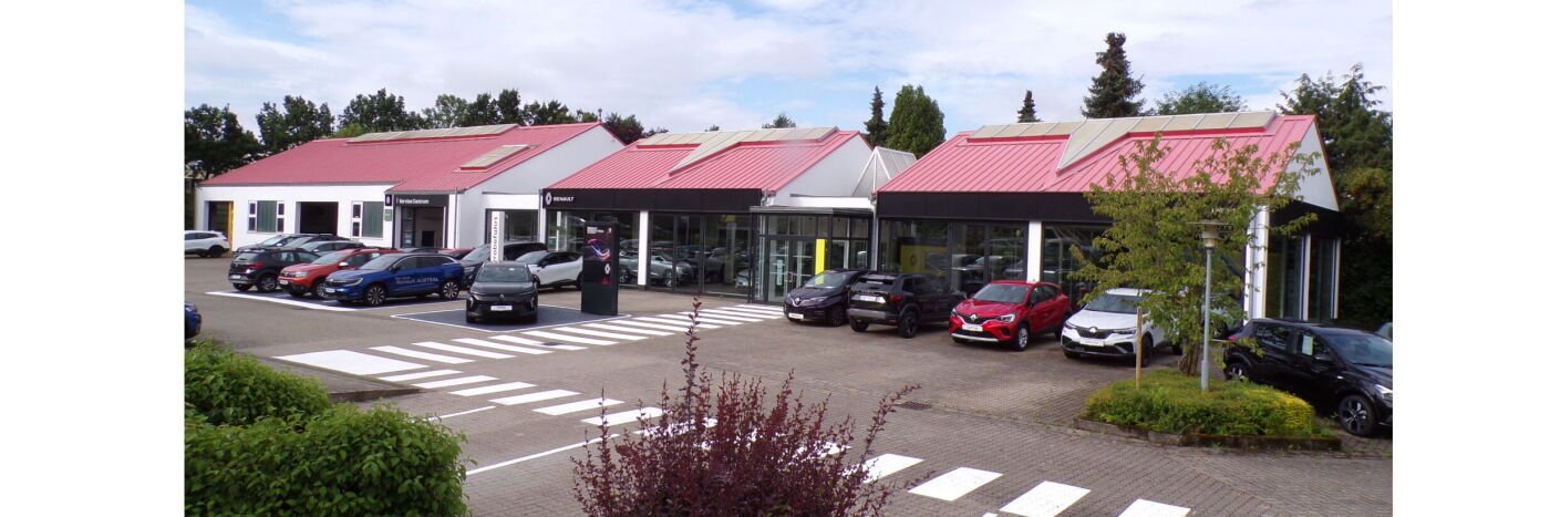 Autohaus mit roten Dächern und Parkplätzen, umgeben von Pflanzen und Bäumen, bei klarem Wetter.