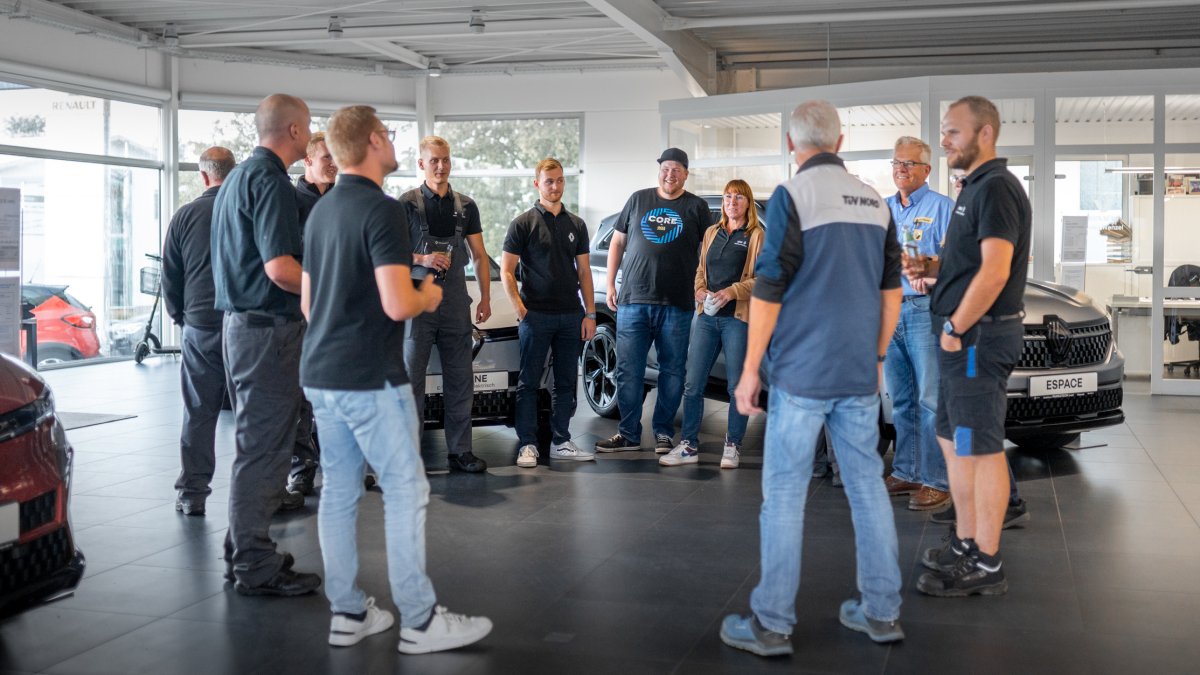 Eine Gruppe von Menschen steht in einem Autohaus und diskutiert, umgeben von Autos.