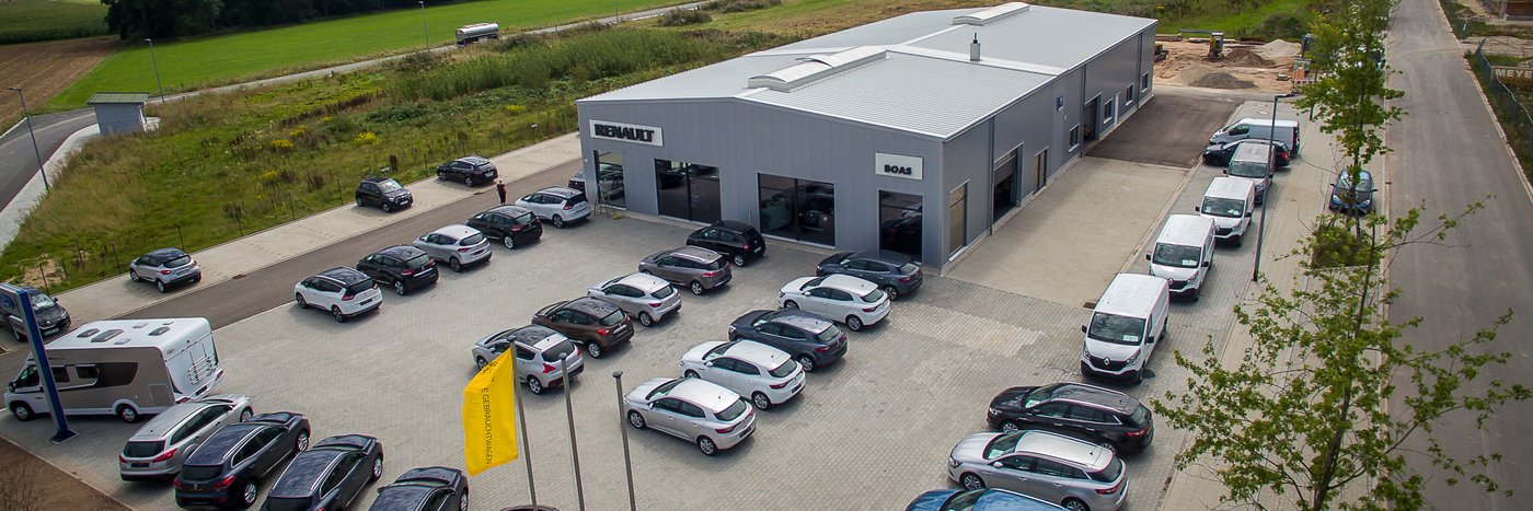 Renault Autohaus mit Parkplatz, umgeben von verschiedenen Fahrzeugen und einer grünen Landschaft.