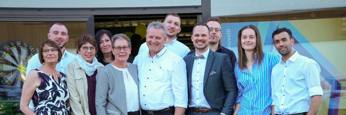 Gruppe von Personen vor einem Geschäft, fröhlich posierend, mit Dartspiel im Hintergrund.