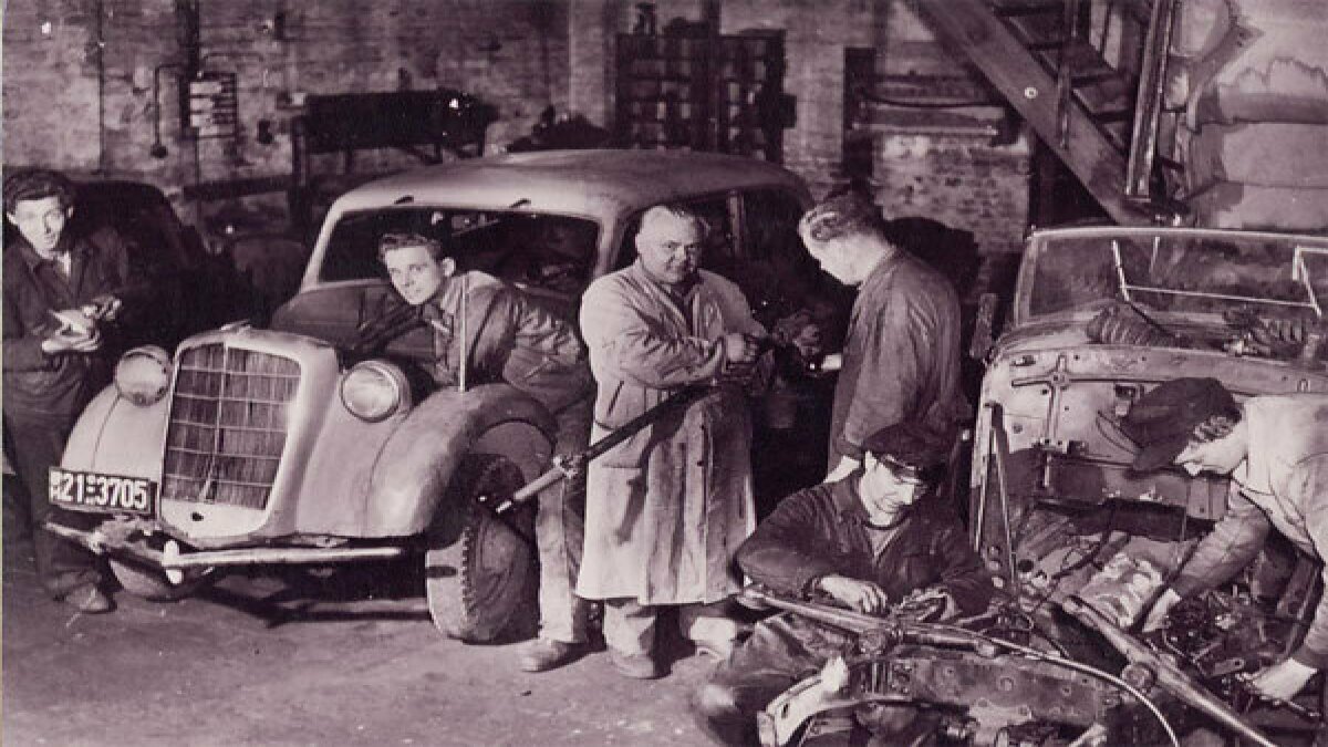 Eine historische Werkstatt mit Mechanikern, die an Autos arbeiten. Alte Fahrzeuge und Werkzeuge sind sichtbar.