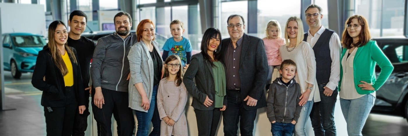Gruppe von Personen, bestehend aus Erwachsenen und Kindern, lächelnd in einem Autohaus. Fahrzeuge im Hintergrund.