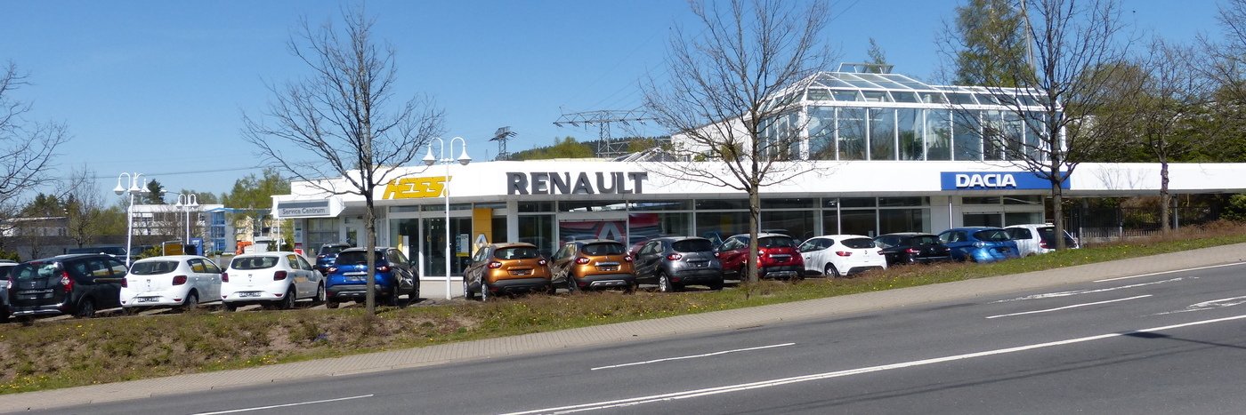 Autohaus mit Renault- und Dacia-Symbolen, PKW auf dem Parkplatz und klaren Himmel. Standort an einer Straßenfront.