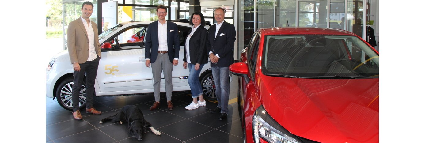 Vier Personen stehen in einem Autohaus neben einem weißen und einem roten Auto, ein Hund liegt auf dem Boden.