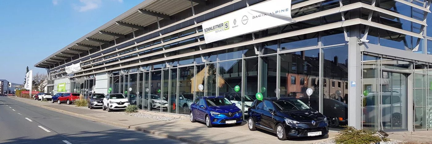 Autohaus mit Glasfassade und mehreren Fahrzeugen vor dem Gebäude, grüne Luftballons dekoriert.