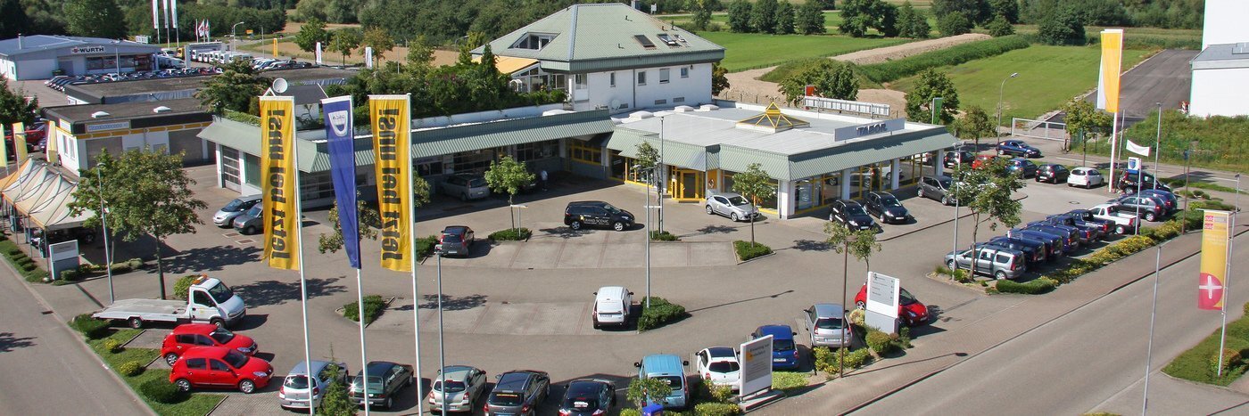 Luftaufnahme eines Autohauses mit Parkplatz und umgebender Landschaft, mehrere Autos und Fahnen sichtbar.