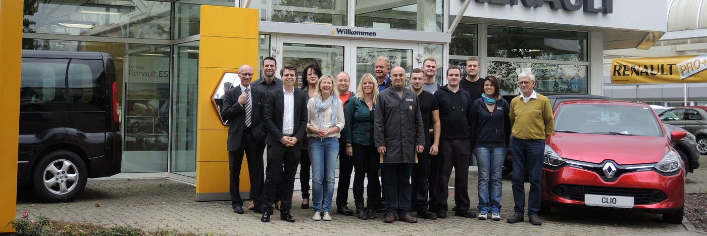 Die Mitarbeiter eines Autohauses stehen vor dem Eingang, neben einem schwarzen und roten Renault.