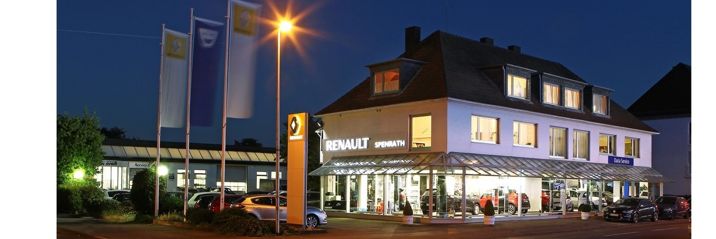 Renault Autohaus Spennath bei Nacht, beleuchtet mit Fahnen vor dem Gebäude und Autos im Showroom.