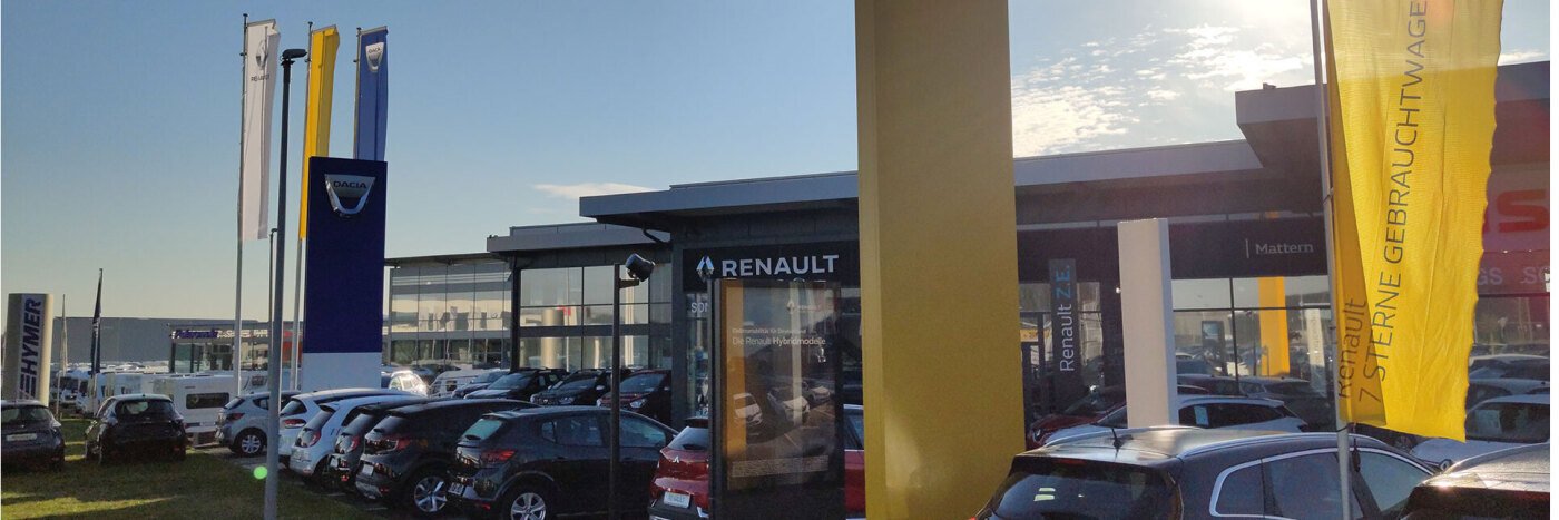 Renault Autohaus mit mehreren Autos auf dem Parkplatz und Flaggen im Vordergrund bei klarem Himmel.