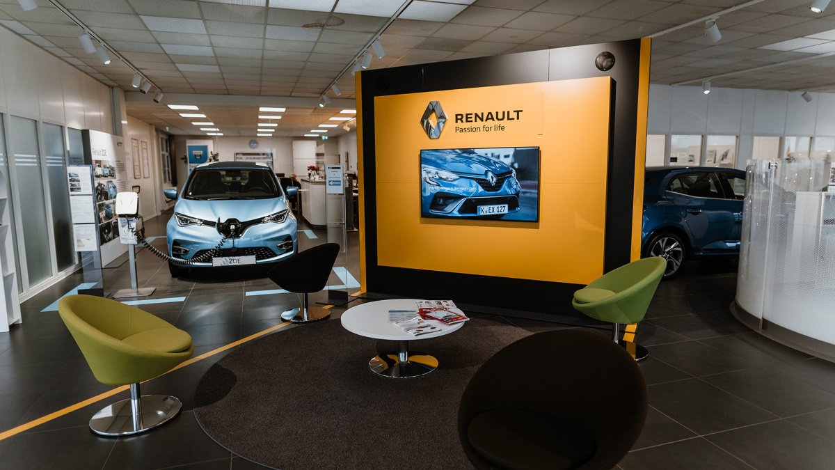 Renault Ausstellungsraum mit Elektroauto, Sitzbereichen und Bildschirm, der Fahrzeuge zeigt.