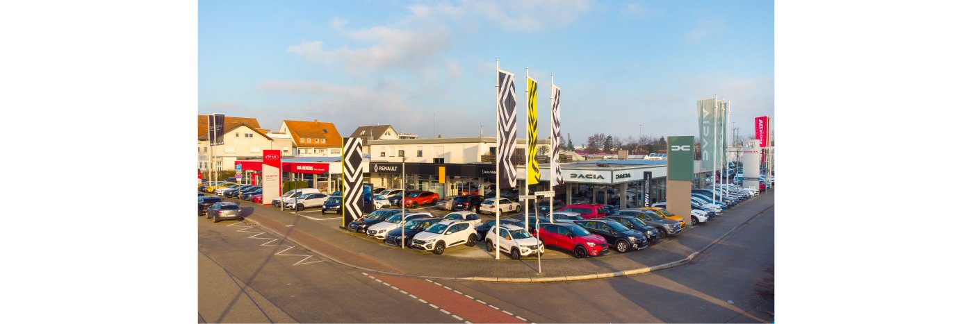 Autohaus mit mehreren Fahrzeugen auf dem Parkplatz, Markenbanner und Gebäude im Hintergrund.