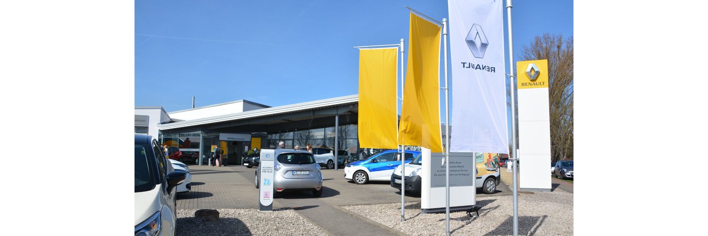 Renault Autohaus mit Flaggen und parkenden Autos an einem sonnigen Tag im Freien.