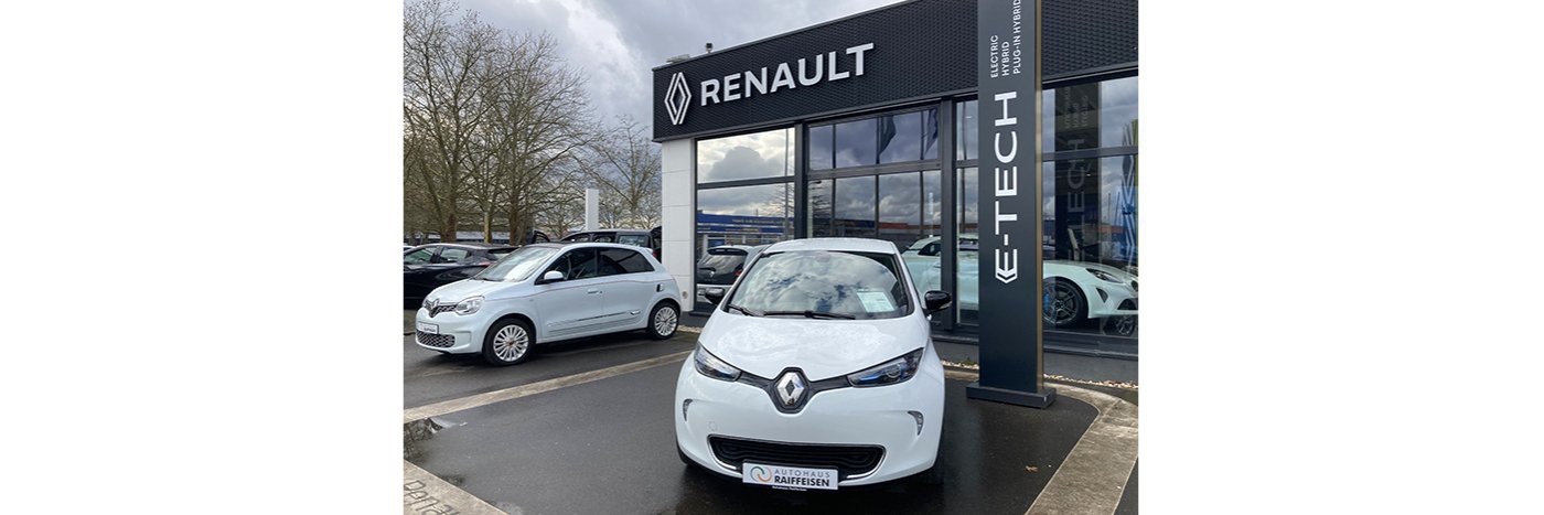 Weißer Renault Zoe vor einem Renault Autohaus mit E-TECH Werbung und mehreren Fahrzeugen im Hintergrund.
