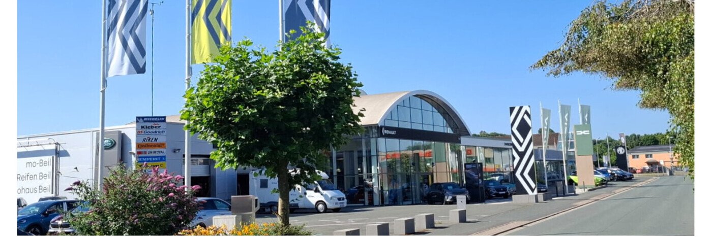 Autohaus mit modernen Flaggen, Fahrzeugen und einer großen Glasfront an einem sonnigen Tag.