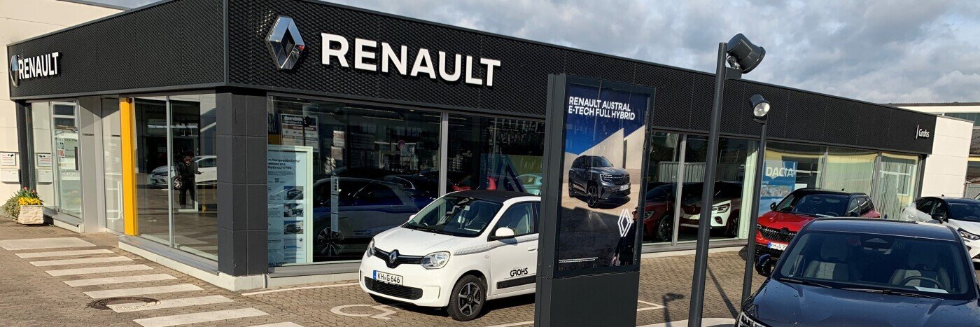 Renault Autohaus mit mehreren Fahrzeugen auf dem Parkplatz und einem Werbeschild für ein Hybridmodell.