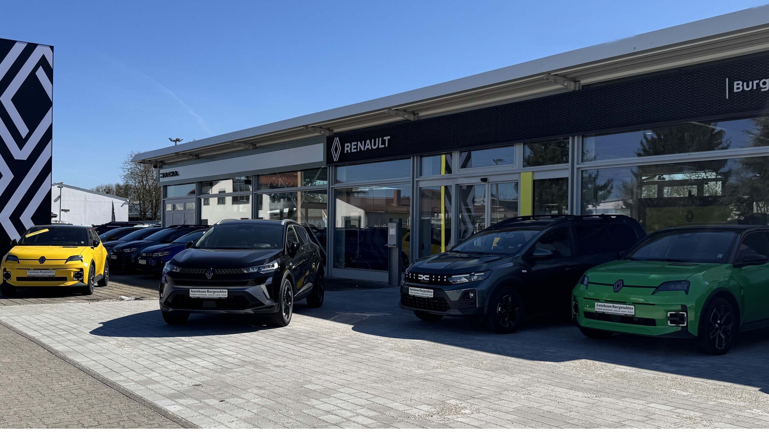 Renault Autohaus mit mehreren Fahrzeugen, darunter schwarze, grüne und gelbe Modelle, vor einem blauen Himmel.