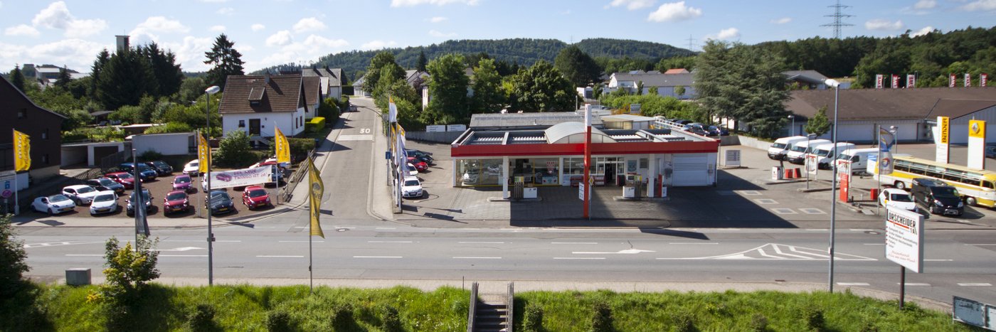 Luftaufnahme eines Parkplatzes mit einer Tankstelle, umgeben von Wohnhäusern und einer grünen Landschaft.