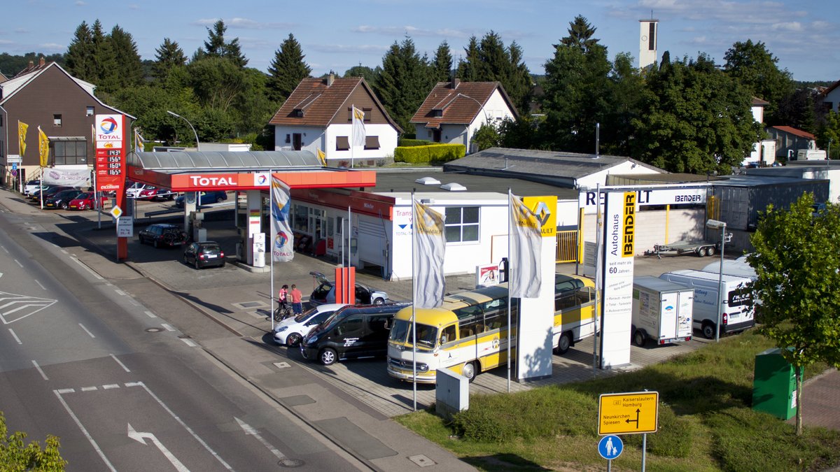 Luftaufnahme einer Tankstelle mit Autos, Flaggen und angrenzenden Wohnhäusern in einer ländlichen Umgebung.