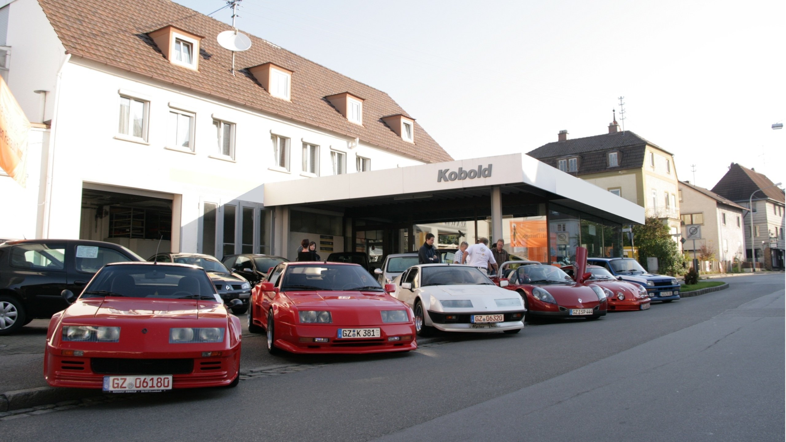 Eine Reihe von Sportwagen steht vor einem Autohaus namens Kobold. Die Autos sind in verschiedenen Farben, hauptsächlich rot.