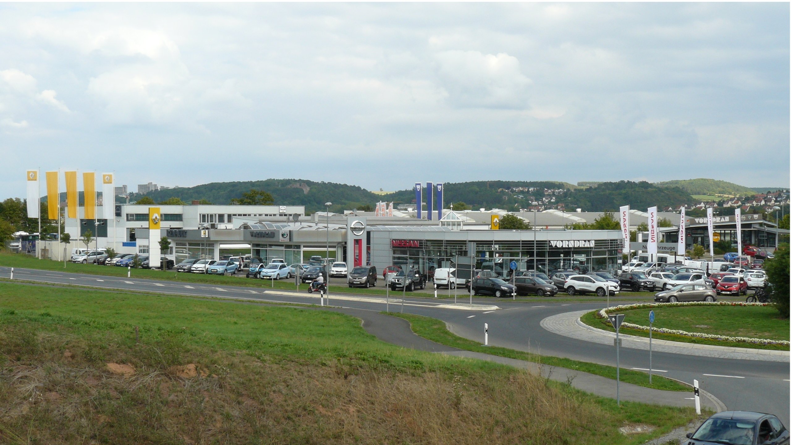 Autohaus und Verkaufsfläche mit Parkplätzen und Autos, umgeben von grüner Landschaft und Hügeln.