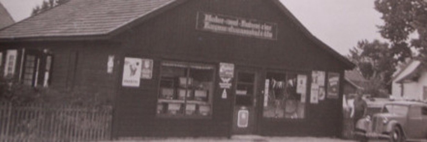 Historisches Ladenlokal mit schwarz-weißer Fassade und Fenstern, umgeben von Bäumen und einem alten Auto.