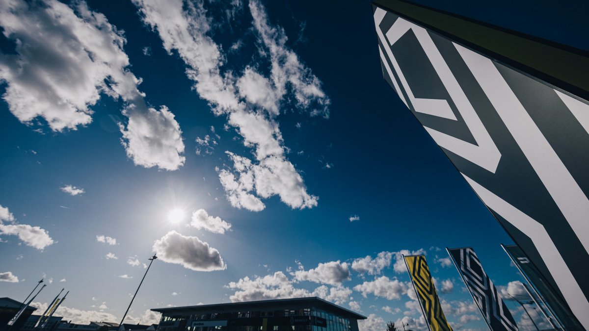 Ein moderner Gebäudekomplex unter einem bewölkten Himmel mit Sonnenstrahlen und grafischen Flaggen im Vordergrund.