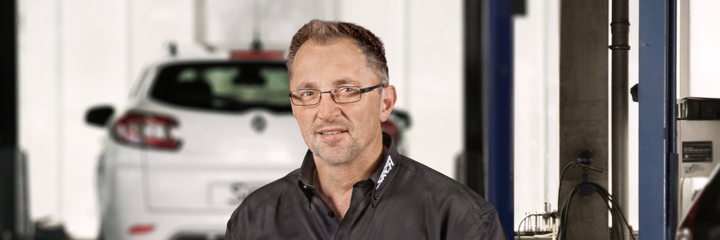Ein Mechaniker in einem Werkstattumfeld, mit einem Auto im Hintergrund. Er trägt eine schwarze Arbeitsjacke und Brille.