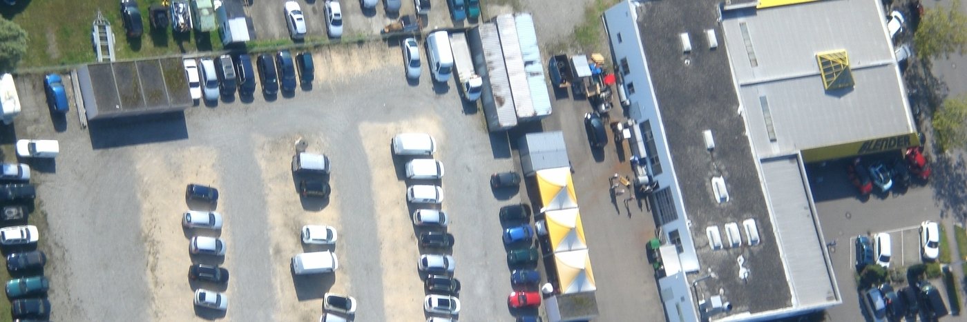 Luftaufnahme eines Parkplatzes mit mehreren Autos und einem Gebäude im Hintergrund. Zubehör und Zelte sichtbar.