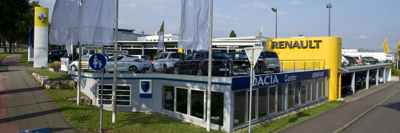 Renault- und Dacia-Autohaus mit Fahrzeugen auf dem Parkplatz, umgeben von Flaggen und Straßenschildern.
