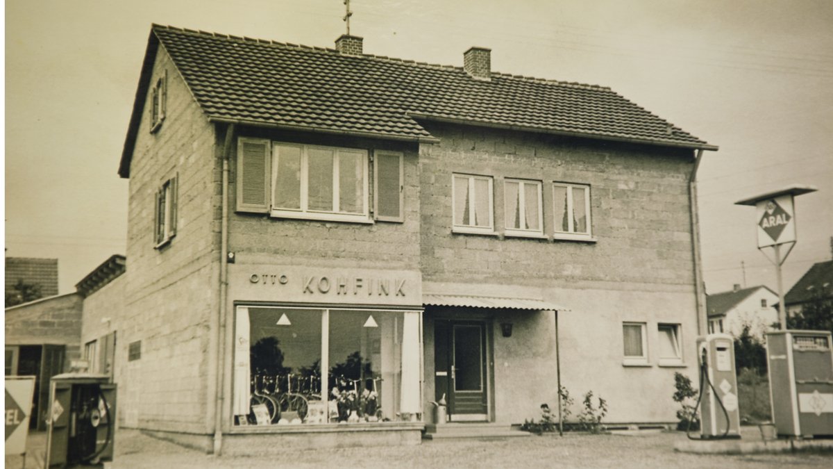 Historisches Foto eines Geschäftsgebäudes mit dem Namen 'Otto Kohfink', an einer Straße mit einer Aral-Tankstelle.