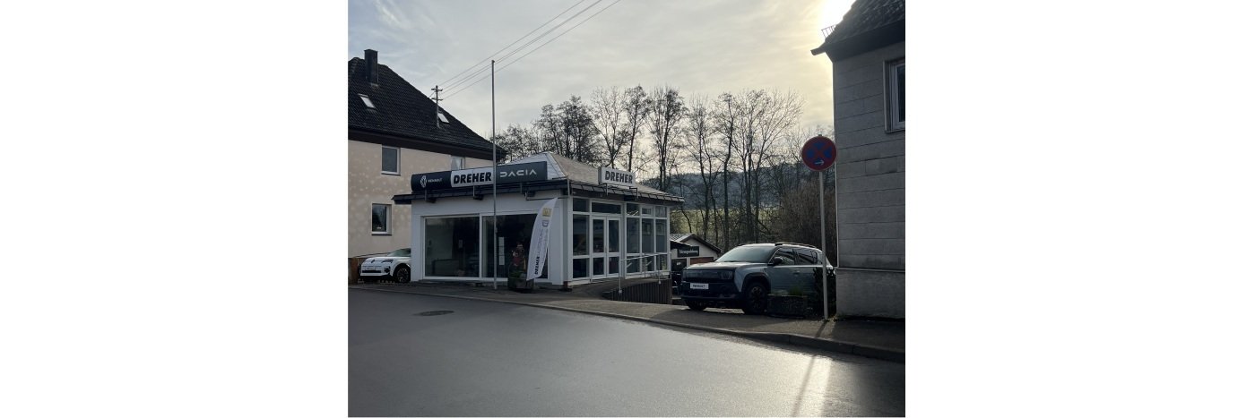 Ein kleines Geschäft mit großen Fenstern, umgeben von Bäumen und Fahrzeugen, in einer ruhigen Straße.