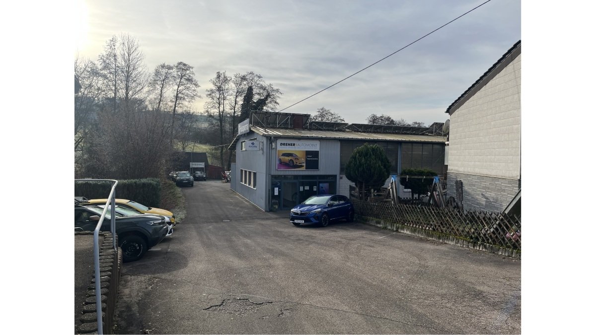 Ein Autohaus mit einer grauen Gebäudeansicht, parkenden Autos und umgebender Natur. Ruhige Straße und bewaldete Hügel im Hintergrund.