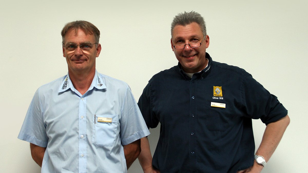 Zwei Männer in Arbeitskleidung stehen zusammen vor einer weißen Wand, lächeln freundlich in die Kamera.